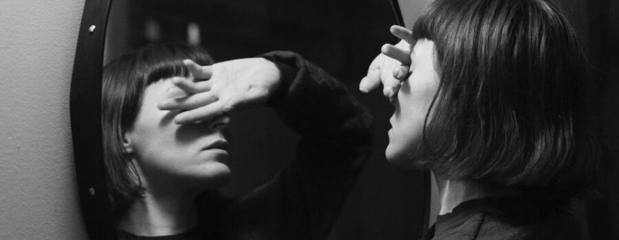 B&W photo of woman with short dark hair standing in front of a mirror with her right hand covering her eyes. Shows both her and her reflection. This photo in on www.becomingstoic.net in a post called "Who Are You?".