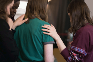 Three women surround a woman wearing a green shirt. They are laying their hands on her shoulder to show support. This photo is on www.becomingstoic.net in a post called "Devotion and Duty".