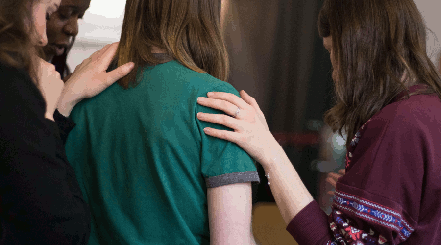 Three women surround a woman wearing a green shirt. They are laying their hands on her shoulder to show support. This photo is on www.becomingstoic.net in a post called "Devotion and Duty".