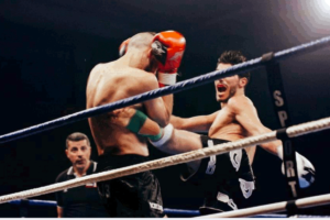 Man on right is throwing a roundhouse kicks to the ribs of his opponent on the left. The referee is standing in the background watching intently. Black background a ropes in the foreground. This photo is used in www.becomingstoic.net in an article called "Stoic Goal Setting".