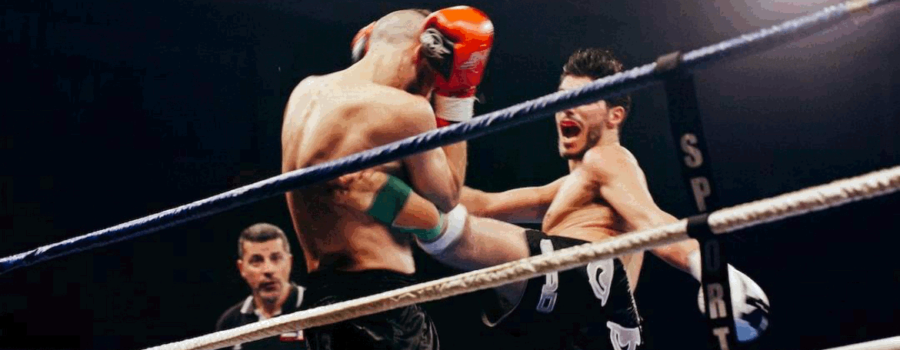 Man on right is throwing a roundhouse kicks to the ribs of his opponent on the left. The referee is standing in the background watching intently. Black background a ropes in the foreground. This photo is used in www.becomingstoic.net in an article called "Stoic Goal Setting".