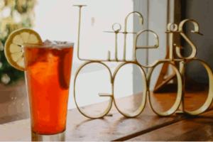 A cool looking glass of tea with a lemon slice on the rim to the left of the table. On the right side of the table is a brass looking statue that is shaped in the words "Life is Good". Focusing on a good life is an important Stoic concept. This photo is on www.becomingstoic.net on a post called "Focusing On A Good Life".