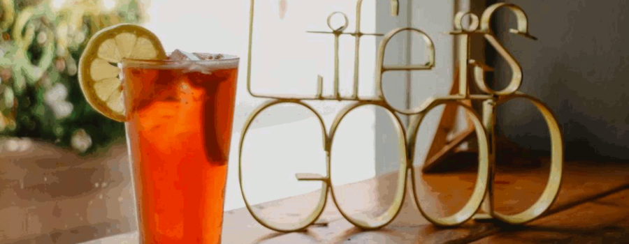 A cool looking glass of tea with a lemon slice on the rim to the left of the table. On the right side of the table is a brass looking statue that is shaped in the words "Life is Good". Focusing on a good life is an important Stoic concept. This photo is on www.becomingstoic.net on a post called "Focusing On A Good Life".