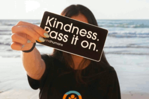 A woman with dark hair stands on a beach facing with her back to the waves. She is holding a black card with white text that states "Kindness Pass it on." This photo is used on www.becomingstoic.net in a post titled "Kindness Isn't Weakness".
