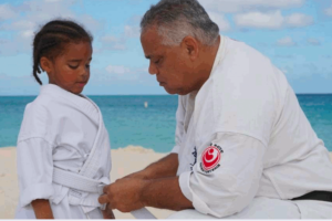 Older male blackbelt kneeling as he ties the belt of young girl with a scene of a beach behind them. This photo is used on www.becomingstoic.net in a post called "Leadership by Default'.