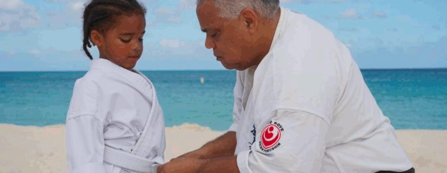 Older male blackbelt kneeling as he ties the belt of young girl with a scene of a beach behind them. This photo is used on www.becomingstoic.net in a post called "Leadership by Default'.